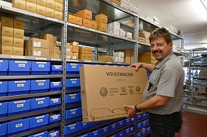 Ein Mitarbeiter in grauem Polohemd steht in einem Ersatzteillager und hält einen großen Karton mit der Aufschrift ‚Volkswagen Group‘, auf dem Logos von Volkswagen, Audi, Seat, Škoda und Volkswagen Nutzfahrzeuge abgebildet sind. Im Hintergrund Regale mit blauen Kisten und braunen Kartons. Der Mann schaut lächelnd in die Kamera.