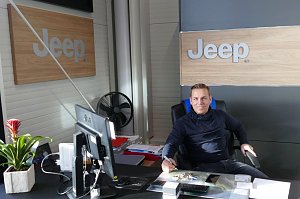Verkaufsberater Danny Kindervater sitzt an einem Schreibtisch in einem modernen Jeep-Autohaus, mit Jeep-Logos an der Wand, Computer und Pflanzen auf dem Tisch.