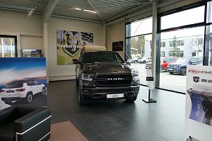 Großer schwarzer RAM-Pickup im Ausstellungsraum eines Autohauses mit großen Fenstern, daneben Werbeplakate für Jeep und Dodge.