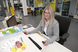 Junge Frau mit langen, blonden Haaren und Brille sitzt am Empfangsschalter des Autohauses. Sie tr&auml;gt einen grauen Cardigan &uuml;ber einer wei&szlig;en Bluse und l&auml;chelt leicht in die Kamera. Auf dem Schreibtisch liegen Prospekte, ein Monitor steht vor ihr. Im Hintergrund sind Verkaufs- und Beratungsbereiche des Opel-Autohauses sichtbar.