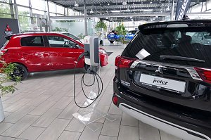 Innenraum eines Autohauses mit mehreren Mitsubishi-Fahrzeugen, im Vordergrund eine Ladestation f&uuml;r Elektrofahrzeuge zwischen einem roten Kleinwagen und einem schwarzen SUV.