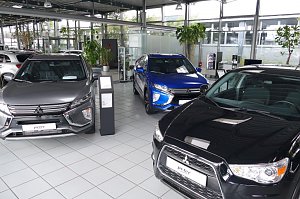 Innenansicht eines Autohauses mit mehreren Mitsubishi-Fahrzeugen in verschiedenen Farben, darunter ein grauer, ein blauer und ein schwarzer Wagen, auf gl&auml;nzendem Fliesenboden mit gro&szlig;en Pflanzen und Glasw&auml;nden im Hintergrund.