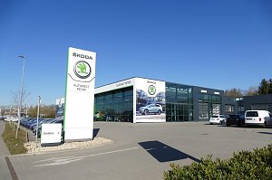 Modernes �koda-Autohaus Autowelt Peter mit gro&szlig;em Glasfassaden-Design, hohem wei&szlig;en �koda-Schild und mehreren geparkten Autos bei blauem Himmel.
