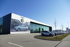 �koda-Autohaus mit gro&szlig;er Glasfassade und gr&uuml;nem Eingang, daneben viele geparkte Autos in einer Reihe, vorne ein gro&szlig;es Werbeplakat mit einem �koda-SUV, blauer Himmel.