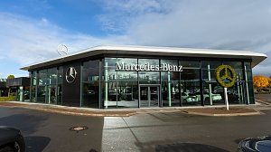 Modernes Mercedes-Benz Autohaus mit gro&szlig;er Glasfassade, Mercedes-Stern-Logo auf dem Dach und bepflanztem Mercedes-Symbol, blauer Himmel mit Wolken.