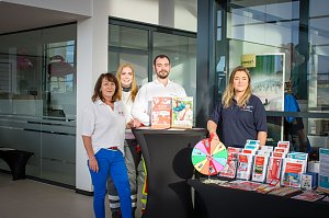 Vier Personen stehen hinter einem Infostand im Škoda Autohaus. Auf dem Tisch liegen Broschüren und ein Glücksrad. Die Personen tragen Kleidung mit Logos des Deutschen Roten Kreuzes.