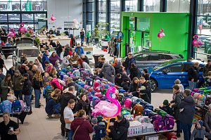 Großer Ausstellungsraum im Škoda Autohaus, gefüllt mit vielen Menschen. In der Mitte sind lange Tische voller bunter Schulranzen. Mehrere Škoda-Fahrzeuge stehen zwischen den Besuchern. Rosa Flamingo-Dekorationen hängen im Raum.