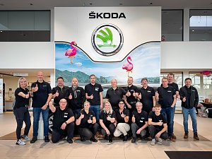 Gruppenfoto von 17 Mitarbeitenden im Škoda Autohaus. Alle tragen schwarze Shirts mit Firmenlogo und zeigen den Daumen nach oben. Im Hintergrund das große Škoda-Logo mit Flamingo-Dekoration.