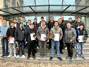 Abschlussklasse mit 16 Auszubildenden sowie einem Ausbildungsleiter und einem Berufsschullehrer auf der Treppe vor dem Eingangsbereich. Alle halten Zeugnisse und Blumen.