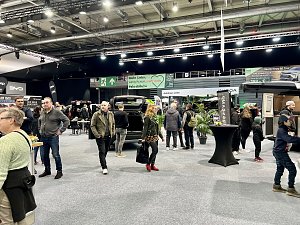 Besucher gehen zwischen verschiedenen St&auml;nden hindurch. Sicht auf SUVs und Camper, mit schwarzen und gr&uuml;nen Messebannern im Hintergrund.