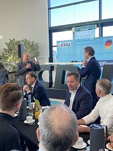 Helmut Peter begr&uuml;&szlig;t mit Mikrofon die G&auml;ste in der Autowelt Peter. Mehrere Personen sitzen an runden Tischen mit Getr&auml;nken und Fr&uuml;hst&uuml;cksgeschirr. Im Hintergrund gro&szlig;e CDU-Buchstaben und eine B&uuml;hne mit Team Merz 2025-Plakat.
