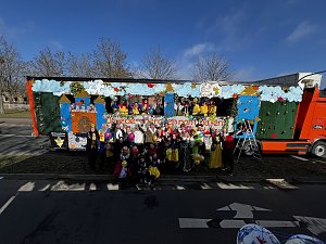 Gro&szlig;e Gruppe verkleideter Menschen posiert vor und auf einem bunt gestalteten Karnevalswagen mit M&auml;rchenmotiven, viele halten bunte Luftballons.