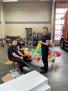 Drei M&auml;nner in schwarzer Arbeitskleidung sitzen und stehen in einer Werkstatt, binden bunte Autohaus-Peter-Luftballons zusammen, im Hintergrund ein orangefarbener LKW.