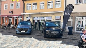 Zwei Mercedes-Transporter – links ein blauer EQV, rechts ein schwarzer Vito – stehen nebeneinander in der Innenstadt von Heiligenstadt. Dazwischen eine schwarze Autohaus-Peter-Flagge, im Hintergrund ein Modegeschäft.