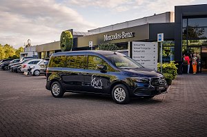 Schwarzer Mercedes-Van mit She�s Mercedes-Logo vor einem Mercedes-Benz Autohaus geparkt. Im Hintergrund stehen weitere Fahrzeuge und einige Menschen am Eingang.