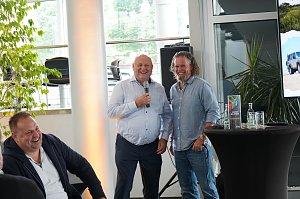 Afrikanischer Abend im Autohaus Peter: Helmut Peter und Abenteuer-Guide Hannu begr&uuml;&szlig;en die G&auml;ste lachend, w&auml;hrend Helmut Peter das Mikrofon h&auml;lt. Links im Bild lacht ein weiterer Teilnehmer, rechts ist ein Stehtisch mit s&uuml;dafrikanischer Dekoration und ein Bildschirm mit einem Safari-Motiv zu sehen.