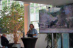 Abenteuer-Guide Hannu beim afrikanischen Abend im Autohaus Peter in Heiligenstadt: Er berichtet am Rednerpult von seinen Safari-Erlebnissen, w&auml;hrend auf einem gro&szlig;en Bildschirm ein Elefant in der afrikanischen Savanne zu sehen ist.