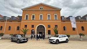 Vorderansicht des historischen Gebäudes mit orangefarbener Fassade. Zwei Mercedes-Benz Fahrzeuge stehen auf dem Platz davor, während sich einige Besucher am Eingang unterhalten. Rechts ist ein Banner mit Werbung für ein kulturelles Event zu sehen.