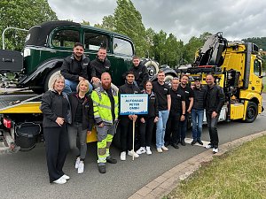 Gruppenfoto eines Teams des Autohaus Peter beim Festumzug in Sondershausen. Die Mitarbeitenden stehen vor einem klassischen Abschleppwagen des ADAC mit einem Oldtimer auf der Ladefl&auml;che. Alle tragen schwarze Autohaus-Peter-Bekleidung, einige lachen in die Kamera. Im Vordergrund wird ein Schild mit der Aufschrift Autohaus Peter GmbH gehalten. Im Hintergrund B&auml;ume und bew&ouml;lkter Himmel.