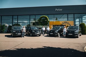 Vor einem modernen Autohaus mit dem Namen Peter stehen vier schwarze Opel-Transporter in einer Reihe. Vier Personen – drei Männer und eine Frau – posieren jeweils neben den Fahrzeugen. An der Glasfassade ist das Opel-Logo und ein gelber Hintergrund zu sehen.