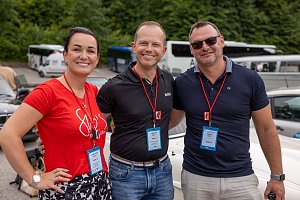 Christina Dost, Carsten Dost und Enrico Dost stehen Arm in Arm vor geparkten Fahrzeugen, tragen Namensschilder und rote Lanyards. Im Hintergrund sind Busse, Autos und grüne Bäume zu sehen – die Szene bei der Rallye wirkt fröhlich.