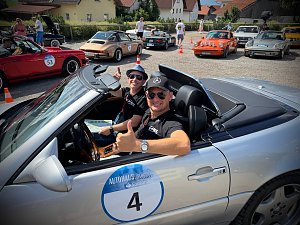 Andreas Peter und Carsten Dost sitzen lächelnd in einem silbernen Mercedes-Benz SL 320 Cabrio mit Startnummer 4 bei einer Oldtimer-Rallye. Beide tragen Sonnenbrillen und zeigen Daumen nach oben. Im Hintergrund stehen mehrere weitere klassische Autos, darunter Porsche-Modelle, auf einem Parkplatz in einer sonnigen Dorfszene. Menschen unterhalten sich und bereiten sich auf die Veranstaltung vor.