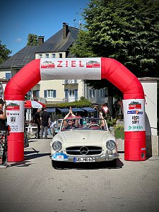 Weißer Mercedes-Benz Oldtimer mit Startnummer 15 fährt unter einem roten Zielbogen mit der Aufschrift ‚ZIEL‘ bei einer Rallye ein. Im Auto jubeln die Insassen mit erhobenen Armen. Menschen stehen rechts und links, klatschen und feiern. Im Hintergrund sind Gebäude mit Balkonen und Bäume zu sehen. Sonniger Tag, festliche Stimmung.