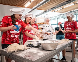 Spielerinnen des Th&uuml;ringer HC in roten und wei&szlig;en Trikots mit wei&szlig;en B&auml;ckerm&uuml;tzen stehen in der Backstube der B&auml;ckerei Helbing. Sie kneten und formen Teig, lachen und haben sichtlich Spa&szlig;. Im Vordergrund liegt Teig auf dem Tisch, daneben eine Sch&uuml;ssel mit Mehl. Eine Frau im Hintergrund filmt die Szene.