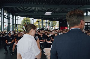 Blick von hinten auf Lilly Hecker und Geschäftsführer Andreas Peter, während sie vor den neuen Auszubildenden sprechen. Viele junge Menschen sitzen in Reihen und hören zu.