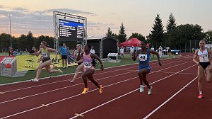 Sprint-Finale über 100 Meter der Frauen. Mehrere Athletinnen im Endspurt kurz vor der Ziellinie, im Hintergrund eine Anzeigetafel mit Namen und Zeiten. Zuschauer und Kampfrichter beobachten den Zieleinlauf