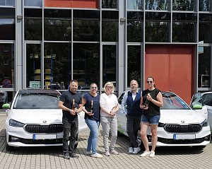 Fünf Personen stehen vor einem modernen Gebäude und posieren mit Champagnerflaschen vor zwei weißen Škoda-Fahrzeugen. Auf den Autos sind grüne Elemente und das Logo der Praxisklinik für Zahnheilkunde zu sehen.