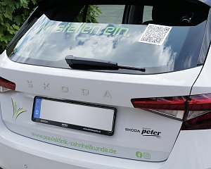 Das Heck eines weißen Škoda-Fahrzeugs mit der Aufschrift Wir stellen ein. und einem QR-Code auf der Heckscheibe. Darunter ist das Logo der Klinik zu sehen.