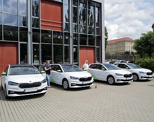 Vier weiße Škoda-Fahrzeuge mit grünen Akzenten stehen nebeneinander vor einem modernen Gebäude. Vor den Autos stehen mehrere Personen mit Flaschen in der Hand, wie bei einer feierlichen Fahrzeugübergabe.