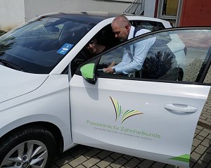 Ein Mann im blauen Sakko zeigt einer Frau auf dem Fahrersitz eines weißen Škoda-Fahrzeugs etwas auf einem Tablet. Auf der Autotür ist das Logo der Praxisklinik für Zahnheilkunde zu sehen.