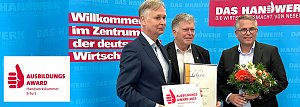 Drei Männer stehen vor einer blauen Wand mit dem Schriftzug „Das Handwerk  Die Wirtschaftsmacht von nebenan“. Einer hält eine Urkunde, ein anderer eine Blumenstrauß, der dritte das Schild „Ausbildungsaward 2025 Handwerkskammer Erfurt“. Das Bild zeigt die Übergabe des Ausbildungsawards an die Autohaus Peter Gruppe.