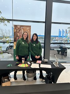 Zwei Frauen in gr&uuml;nen Hoodies stehen an einem Infotisch mit Stiften und Flyern, dahinter ein AOK-Plus-Banner. 