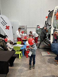 Kinder basteln an einem Tisch mit Rotkreuz-Ballons, daneben Sanit&auml;ter und ein offener Rettungswagen im Autohaus.