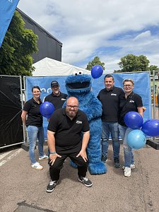 Man sieht 5 Mitarbeiter des Autohaus Peter zusammen mit dem Krümelmonster aus der Sesamstraße mit blauen Luftballons vor einer blauen Bauzaunplane.