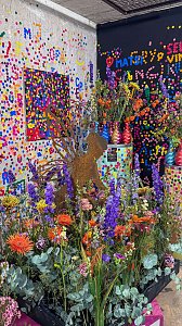Eine farbenfrohe Dekoration mit &uuml;ppigen, bunten Blumenarrangements im Vordergrund vor W&auml;nden, die mit bunten Punkten und abstrakten Mustern im Pop-Art-Stil beklebt sind.