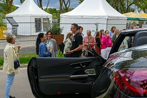 Eine Menschengruppe im Freien, die sich im Hintergrund unterh&auml;lt, w&auml;hrend im Vordergrund die ge&ouml;ffnete Fahrert&uuml;r eines dunklen Mercedes-Cabriolets zu sehen ist.