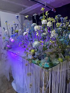 &Uuml;ppige florale Dekorationen aus blau-wei&szlig;en Blumen, die unter k&uuml;hlem, lila-blauem Licht auf einem mit Fransen verzierten Tisch arrangiert sind.
