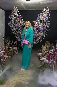 Eine Frau in einem t&uuml;rkisfarbenen Hosenanzug, die l&auml;chelnd vor einer Wand mit gro&szlig;en, kunstvoll aus Blumen geformten Engelsfl&uuml;geln posiert.