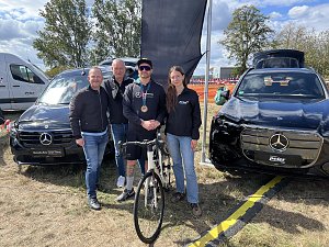 Vier Mitarbeiter des Autohauses Peter stehen f&uuml;r ein Gruppenbild auf einer Wiese zwischen zwei schwarzen Mercedes-Fahrzeugen. Der Mann in der Mitte tr&auml;gt eine Medaille und h&auml;lt ein Rennrad. Im Hintergrund weht eine gro&szlig;e schwarze Fahne.