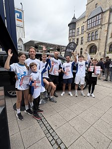 Man sieht unser Firmenlauf-Team nach dem Lauf, vor einem schwarzen EQT. Dahinter steht eine schwarze Beachflag mit unserem Logo in Wei&szlig; und dem wei&szlig;en Mercedes-Benz Stern. Im Hintergrund sieht man das Dessauer Rathaus.