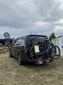 Eine schwarze Mercedes-Benz V-Klasse mit Fahrradtr&auml;ger, auf dem zwei E-Bikes befestigt sind. Im Hintergrund das Festgel&auml;nde.