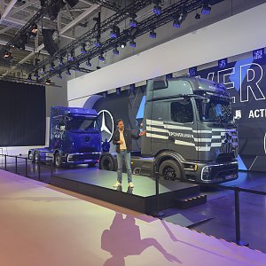 Zwei moderne Mercedes-Benz Lkws auf einer Messeb&uuml;hne. Ein dunkelblauer Lkw links im Hintergrund, ein grau-gestreifter rechts im Vordergrund. Ein Mann steht in der Mitte auf einem Podest und pr&auml;sentiert die Fahrzeuge vor einer gro&szlig;en LED-Wand.