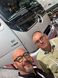 Selfie von zwei M&auml;nnern mit Brille auf einer Messe. Im Hintergrund steht die silberne Front eines Mercedes-Benz Lkws mit gro&szlig;em Logo. Der Mann vorne l&auml;chelt gl&uuml;cklich, der Mann dahinter schaut ruhig in die Kamera.