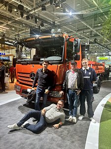 Vier M&auml;nner posieren gl&uuml;cklich vor einem gro&szlig;en, orangefarbenen Mercedes-Benz Lkw auf einer Messe. Drei M&auml;nner stehen aufrecht neben der Sto&szlig;stange, w&auml;hrend der vierte Mann entspannt auf dem grauen Teppichboden davor liegt.