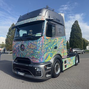 Bunt bemalter Mercedes-Benz Lkw von vorne links auf einem Pflasterplatz. Die Karosserie ist mit wilden, farbenfrohen Mustern bedeckt. Das Dach und die Sto&szlig;stange sind mattgrau. Im Hintergrund blauer Himmel.