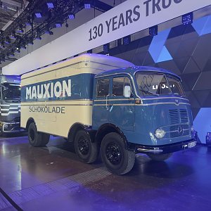 Historischer Mercedes-Benz Lkw auf einer Messe. Blaues Fahrerhaus und cremewei&szlig;er Kofferaufbau mit der Aufschrift MAUXION SCHOKOLADE. Drei Achsen mit schwarzen Felgen. Im Hintergrund h&auml;ngt ein Banner mit 130 YEARS TRUCKS.